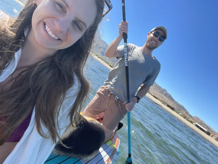 Angela smiling in a selfie while Colin fishes from a paddleboard behind her