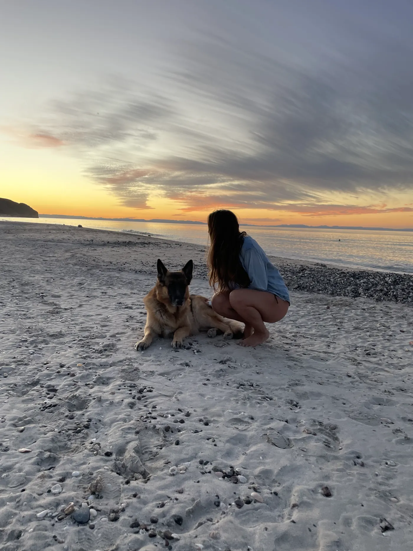 Angela crouching on the beach with Jack, our German Shepherd, under a cloudy sunset