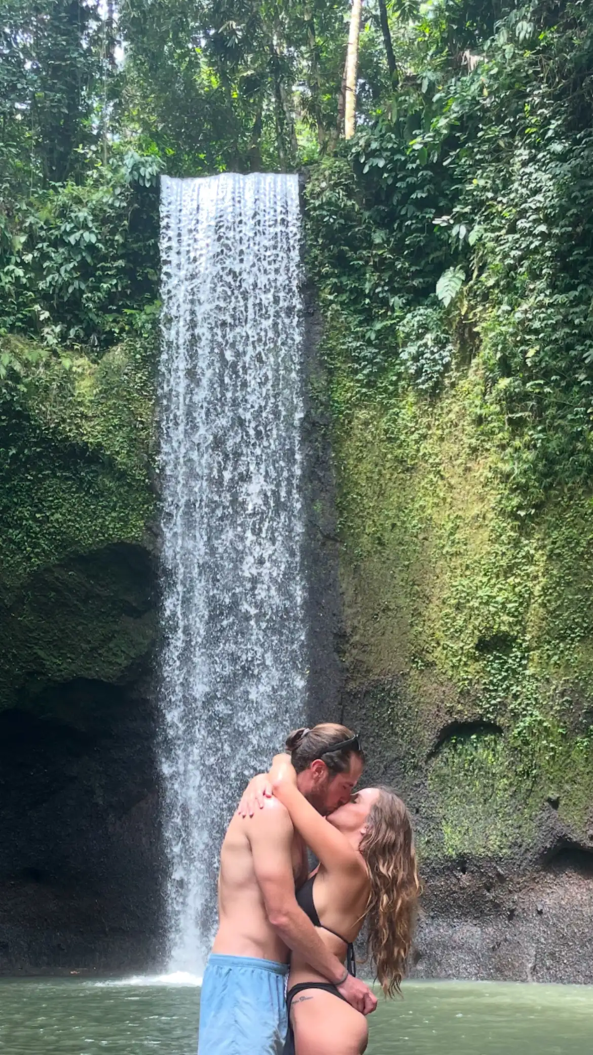 Colin and Angela kissing in front of a tall waterfall in the jungle