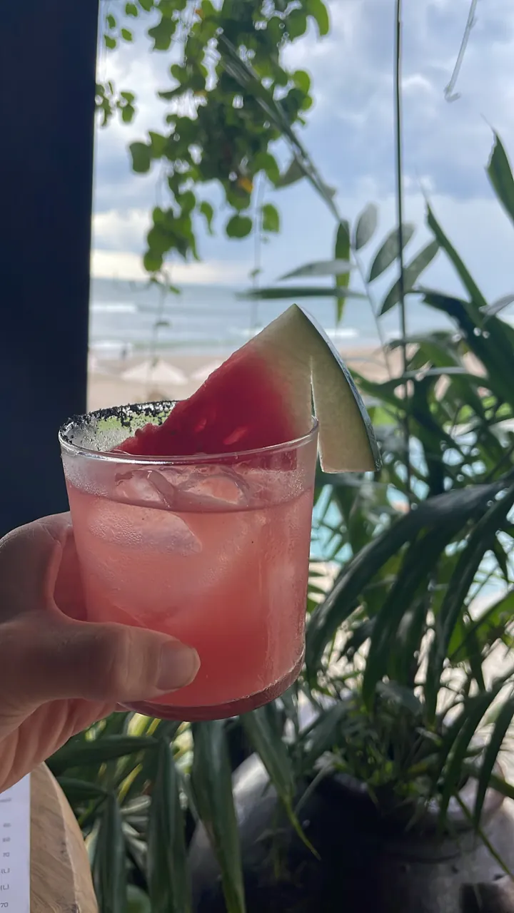 Pink watermelon cocktail garnished with a watermelon slice, held up against tropical greenery