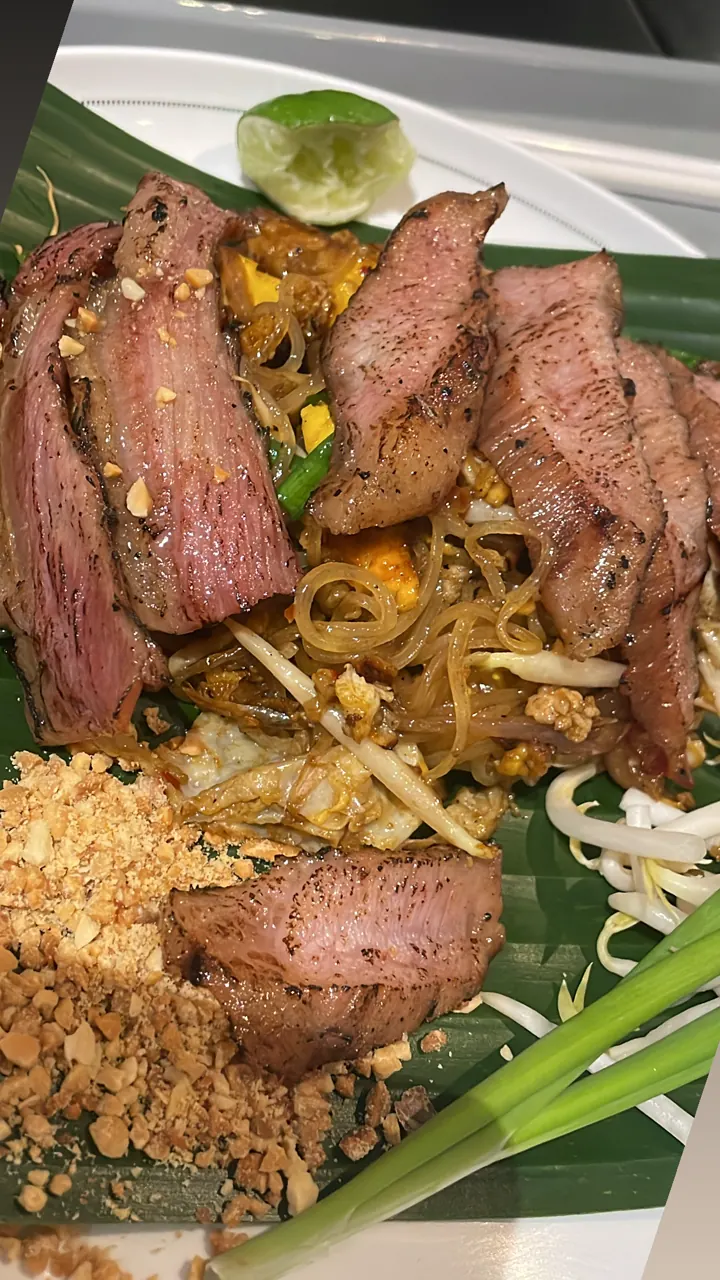 Plate of stir-fried noodles topped with sliced grilled beef and herbs