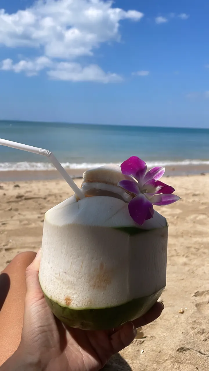 Hand holding a fresh coconut with a straw and pink flower on the beach