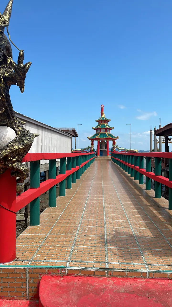 Long wooden pier with red railings leading to a Chinese-style pavilion under blue sky