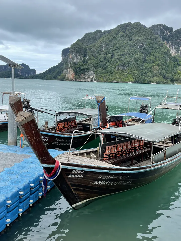 Wooden longtail boats tied up at a floating dock below green limestone hills