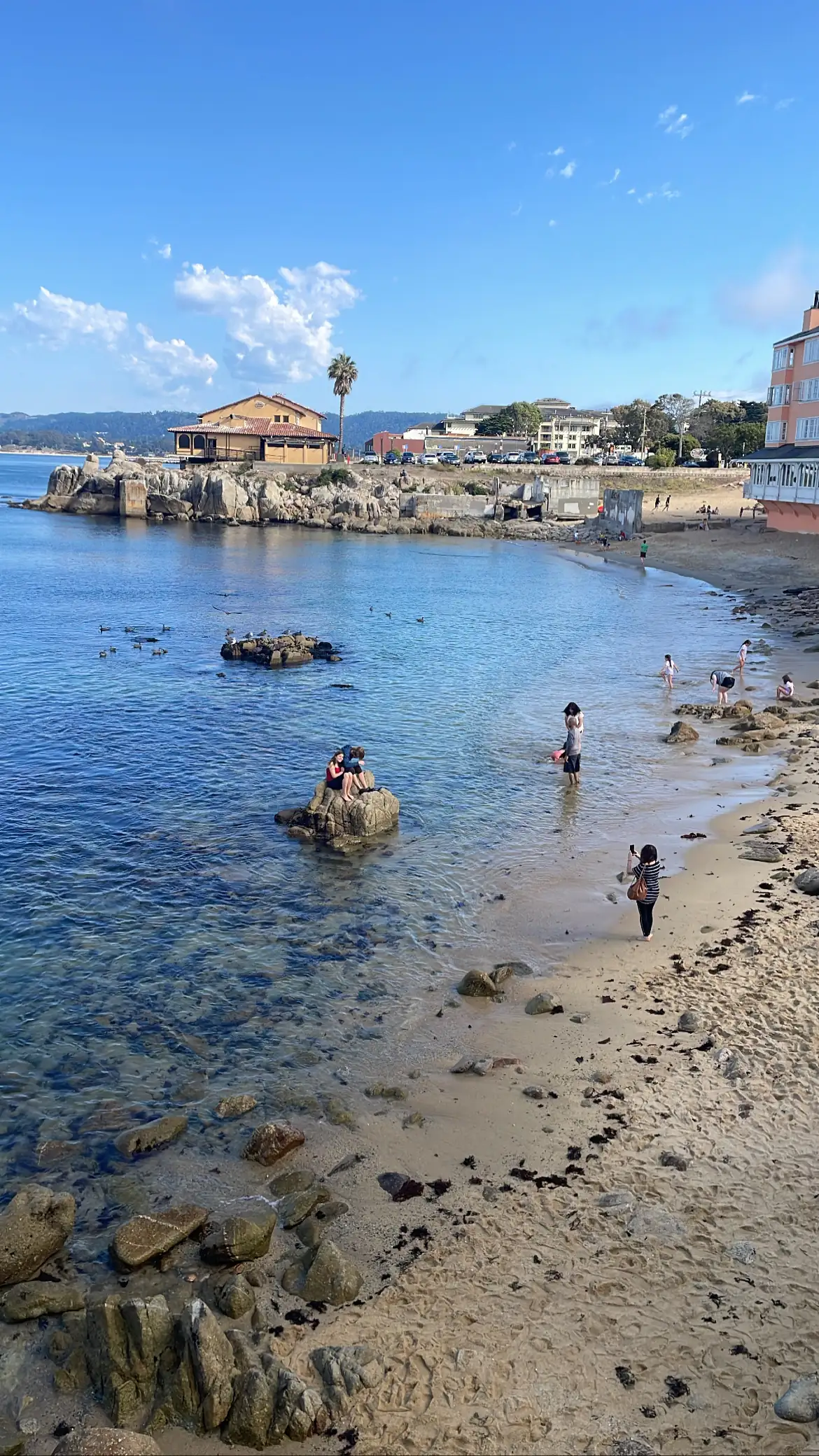 A calm, clear cove with a sandy beach and rocky shore and people wading