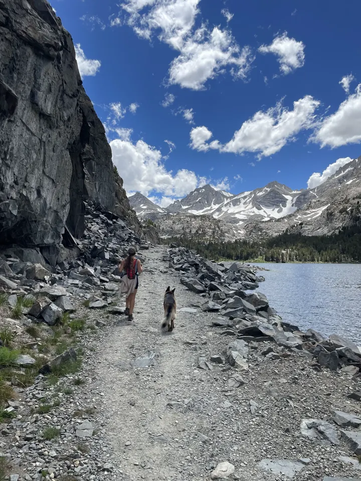 Hikers on a rocky lakeshore trail beneath granite cliffs and puffy clouds