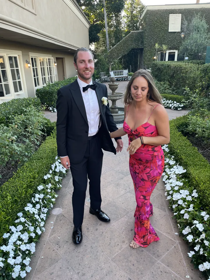 Colin in a tux and Angela in a pink floral gown on a garden path lined with white flowers