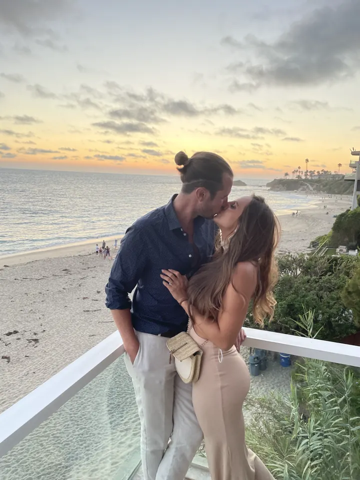 Colin and Angela kissing at a glass railing over the beach at sunset