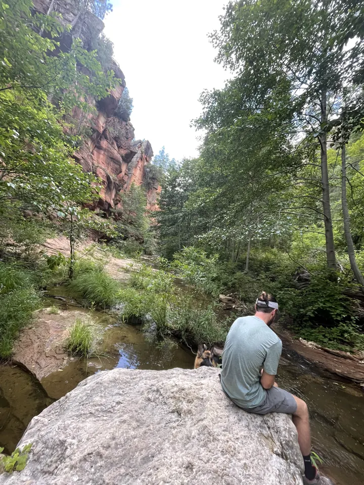Colin sitting on a large boulder above a creek, with red cliffs and green trees