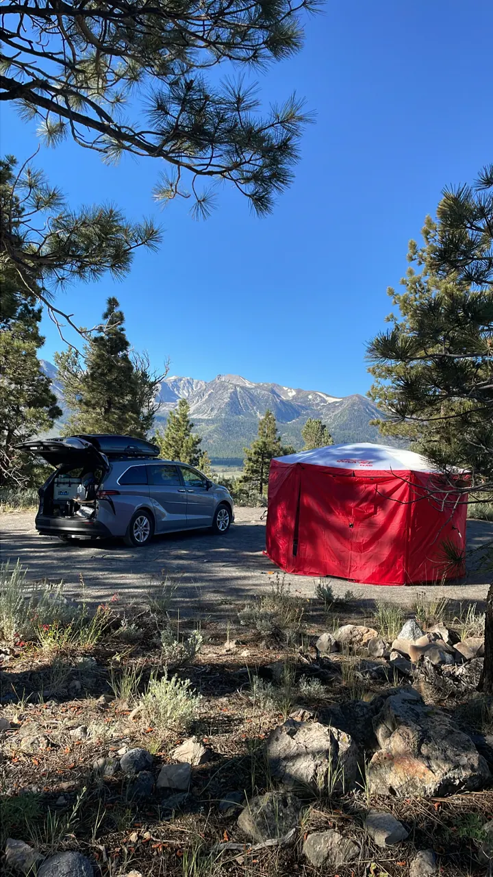 A red tent pitched next to a car under tall pines on a sunny day
