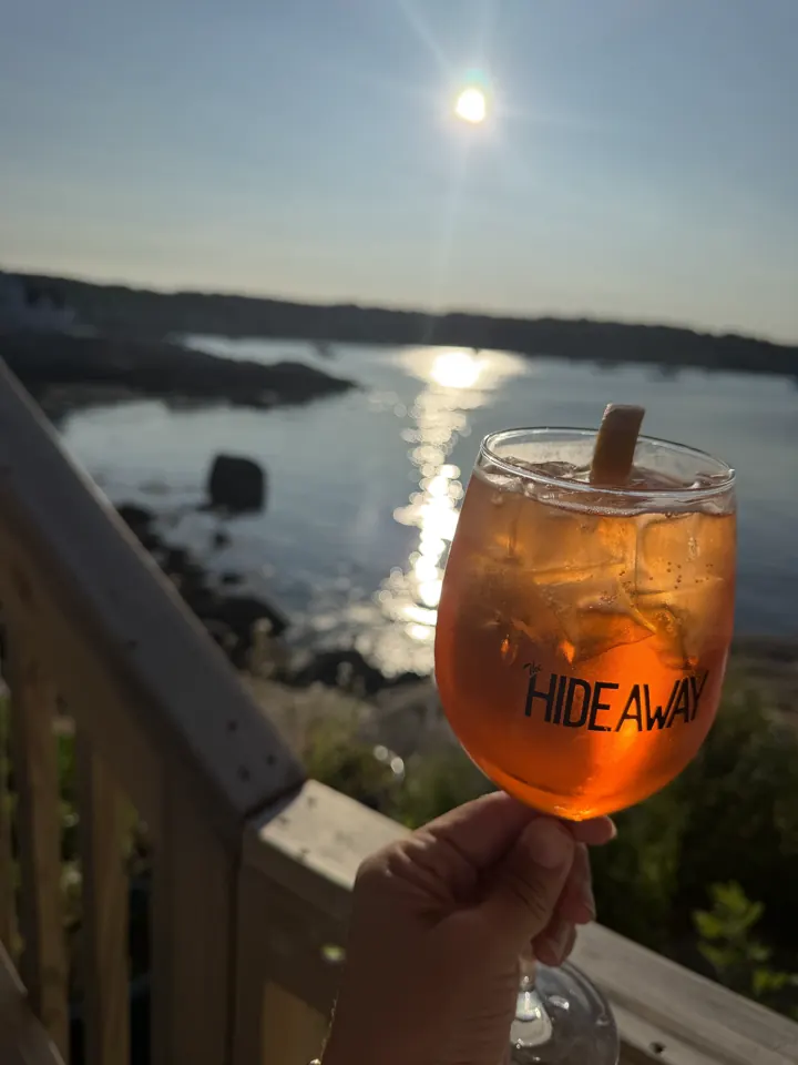 An Aperol spritz held up against a sunset over the water in Rockport