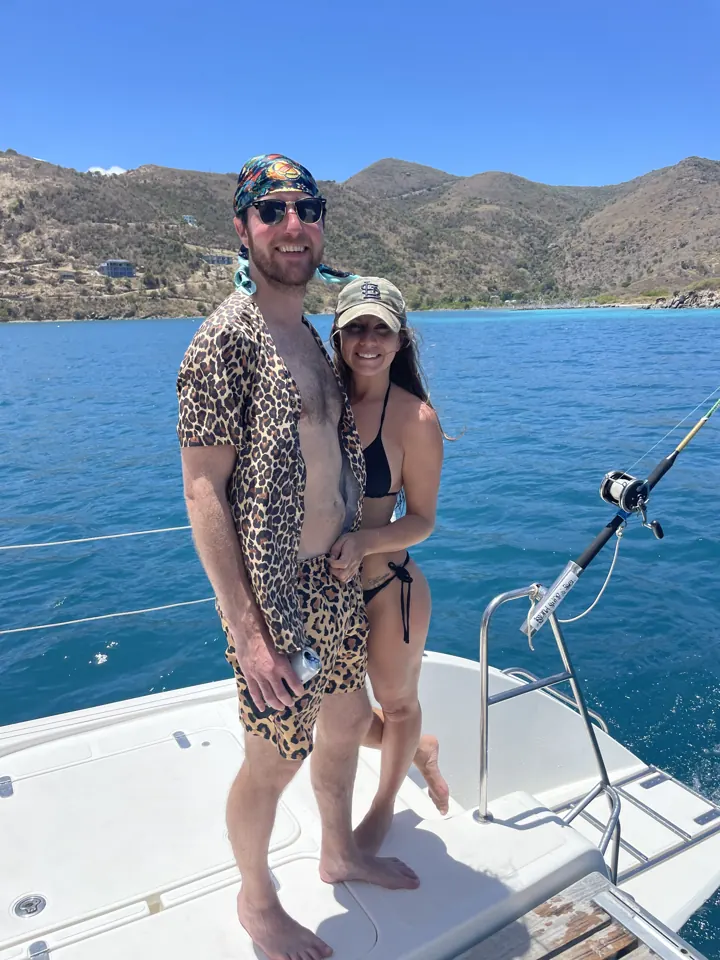 A couple standing on a catamaran with green hills and blue water behind