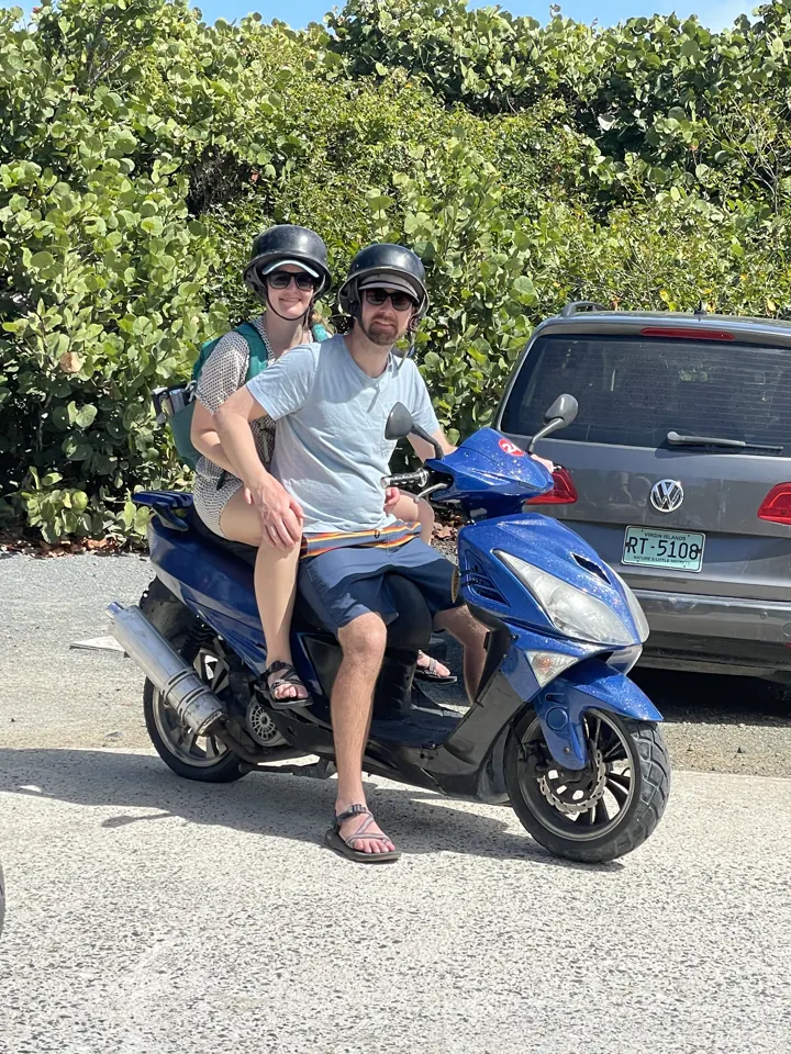 A couple sitting on a blue scooter wearing helmets beside a parked car
