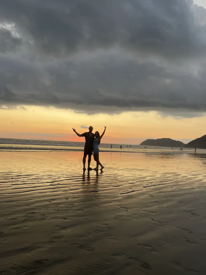 Two people silhouetted with arms raised on a wet beach under a stormy sunset sky