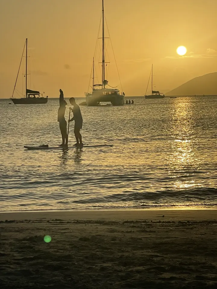 Two people silhouetted in shallow water at sunset with sailboats behind