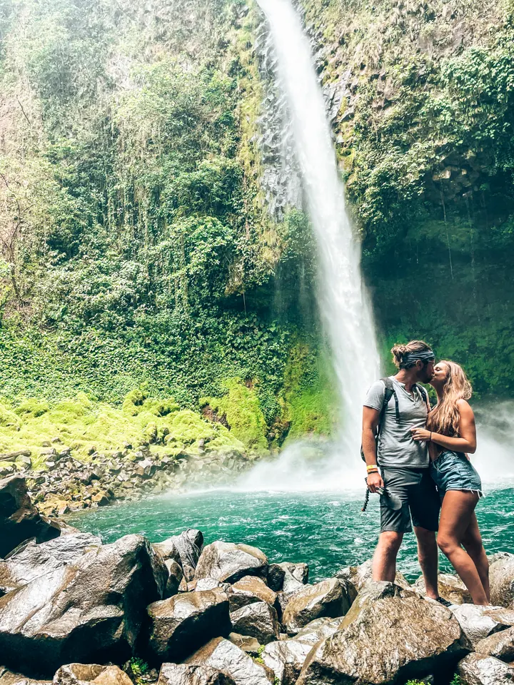 A couple posing on rocks in front of a tall waterfall and turquoise pool