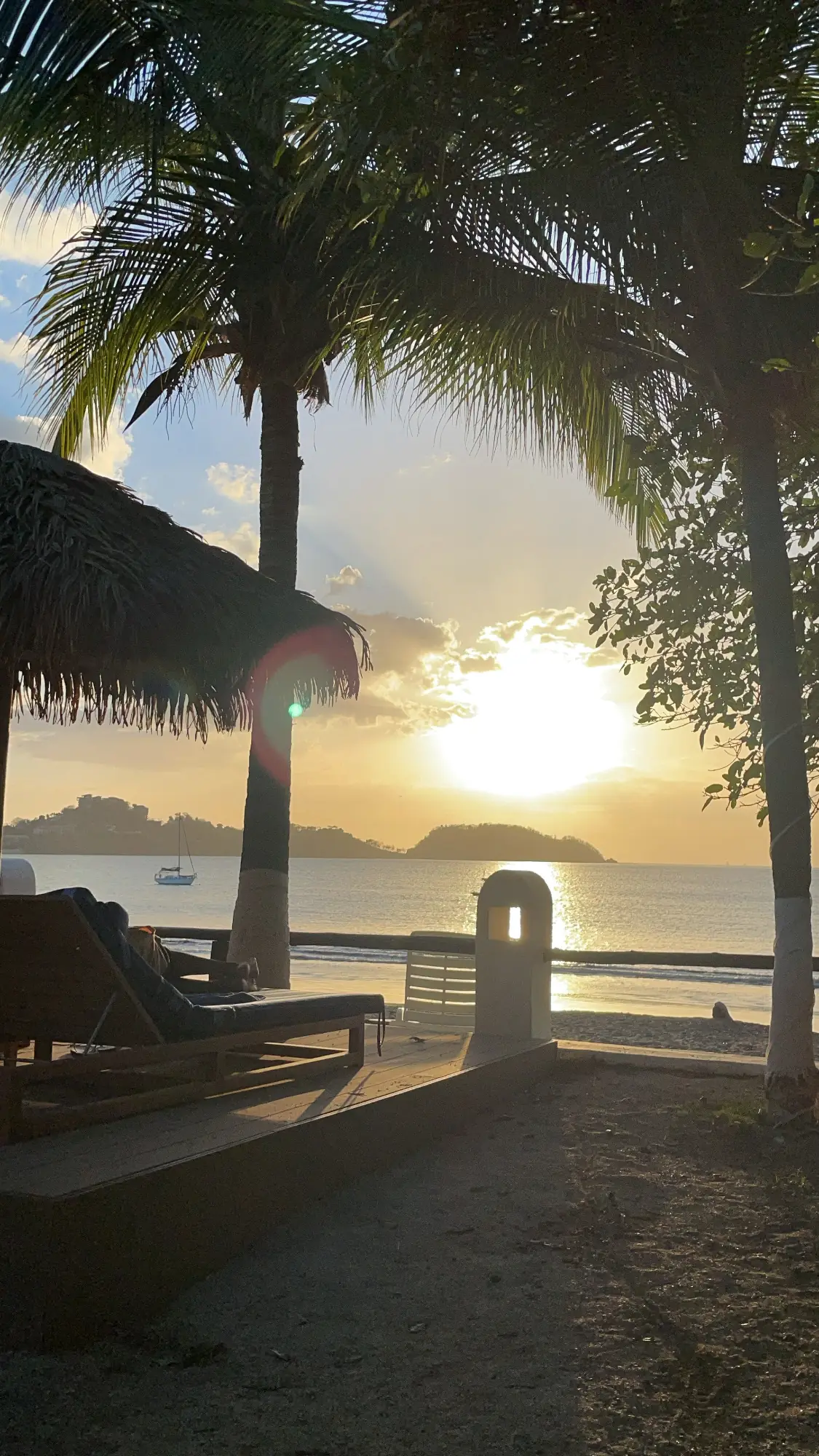 A golden sunset over the Pacific framed by palm trees and a thatched palapa