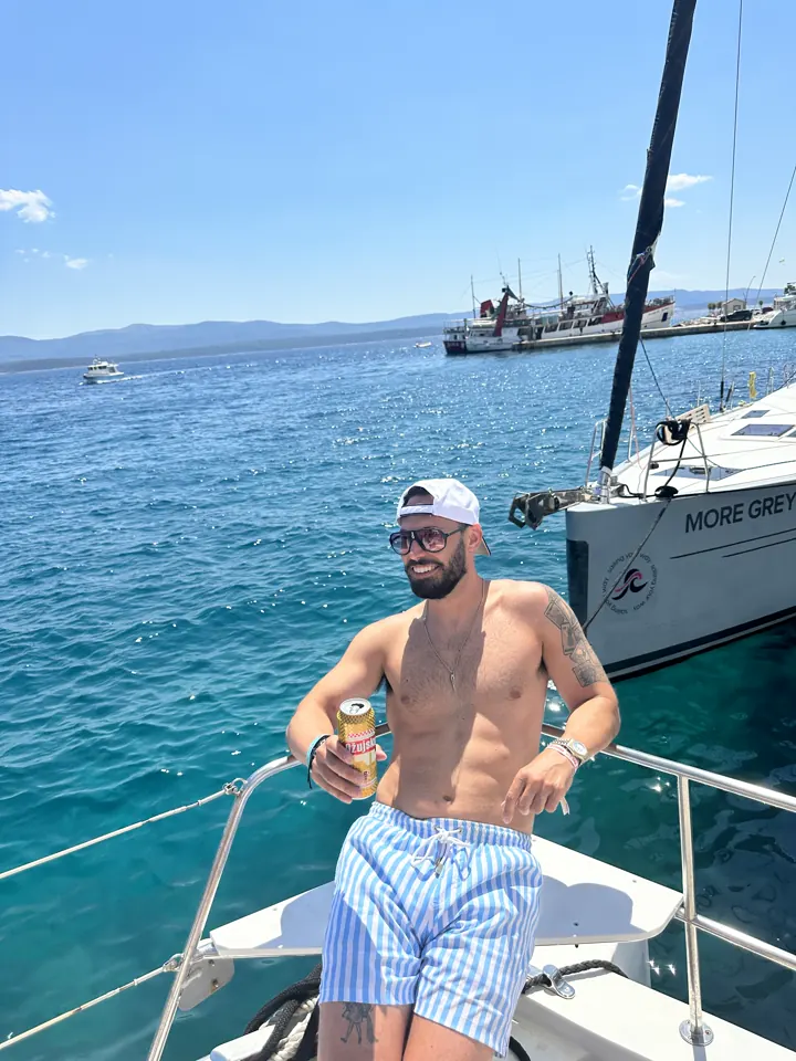 Colin sitting on the bow rail of a boat on bright blue water with a sailboat nearby