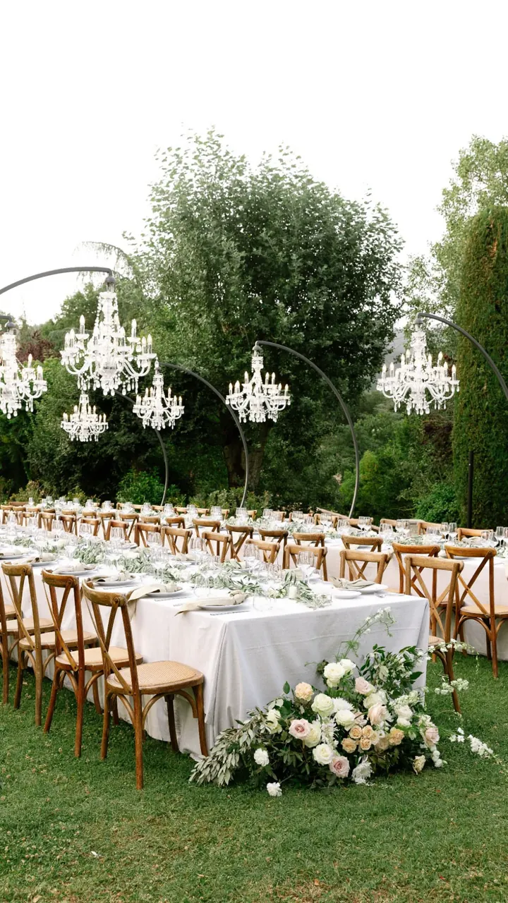 Long white tables with flowers and wooden chairs set on a lawn beneath trees
