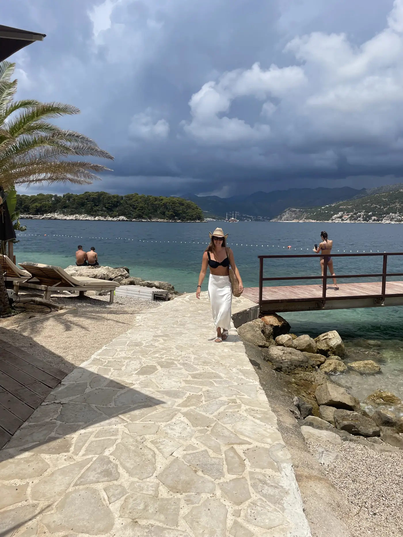 Woman walking along a stone seaside promenade toward a pier, with green islands and blue sea under clouds