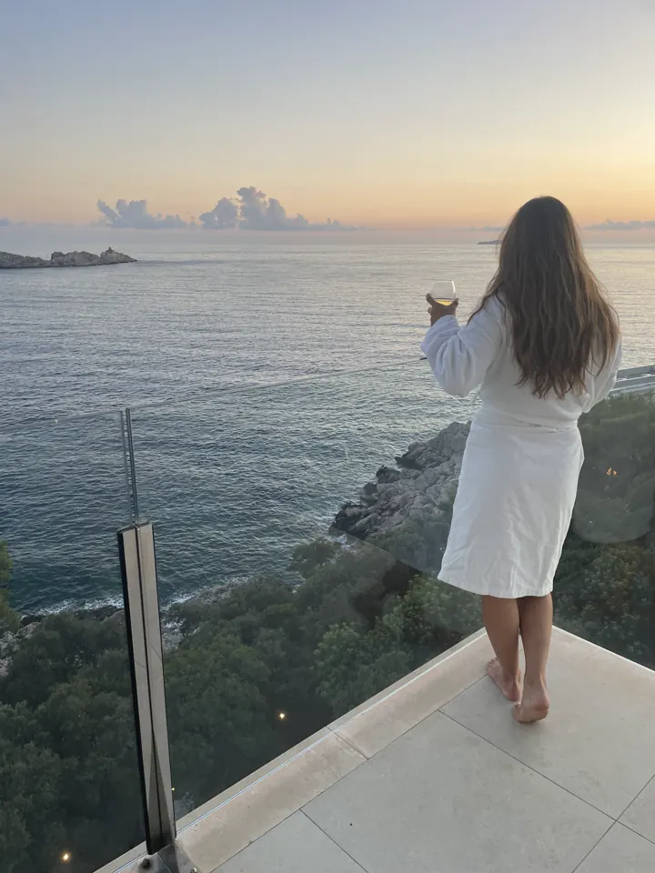 Woman in a white robe standing on a terrace railing looking out over the sea at sunset