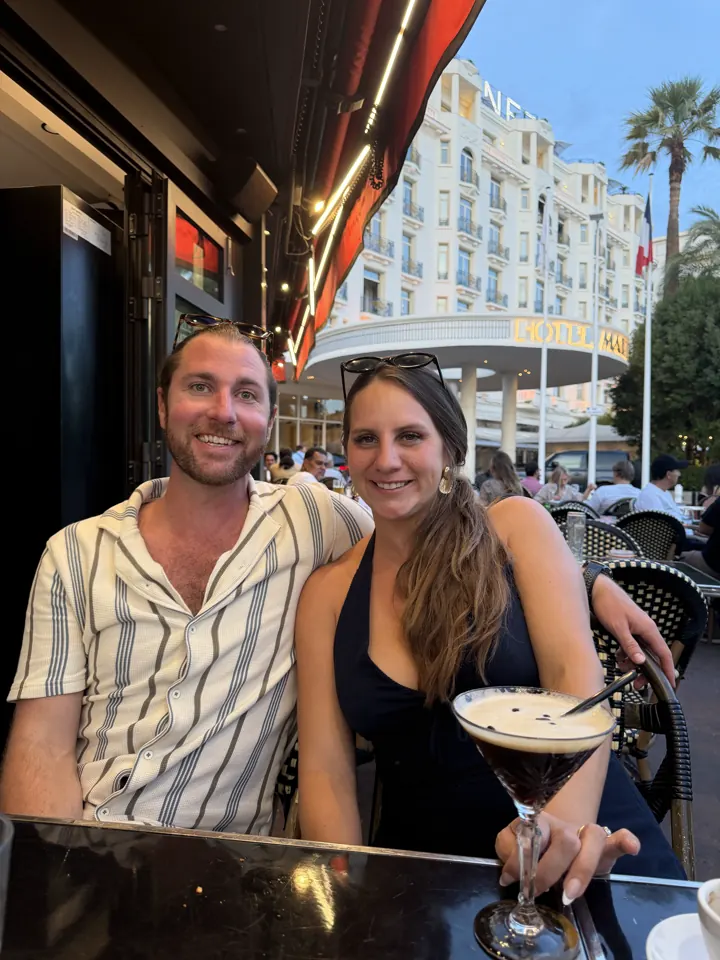 Colin and Angela smiling at a cafe table with drinks, palm trees and a white building behind
