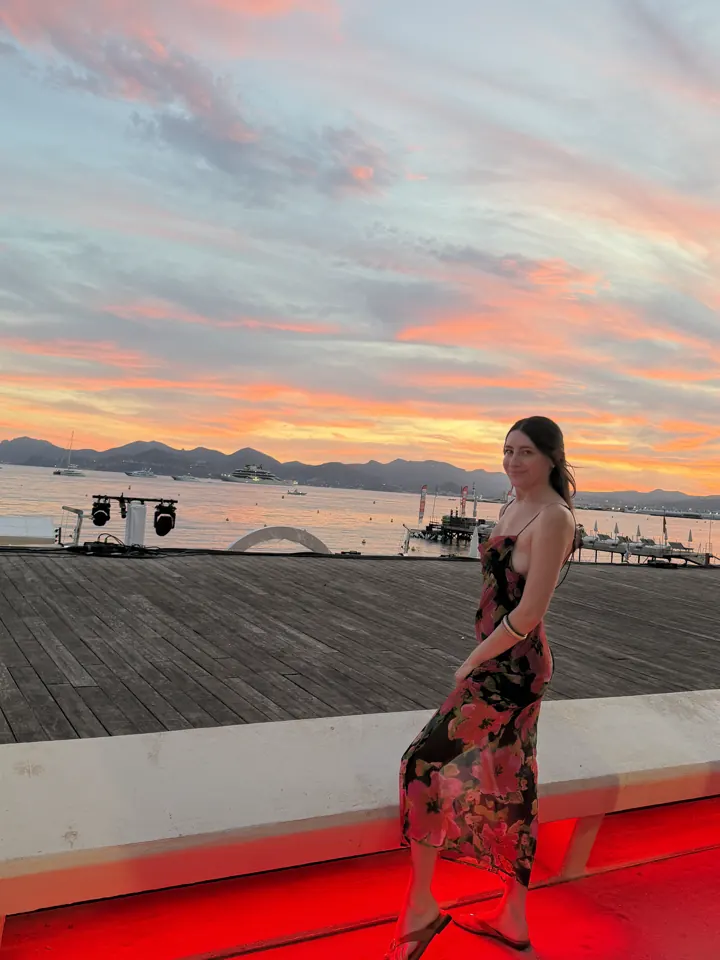 Angela in a floral dress posing by the water under a pink sunset sky in Cannes