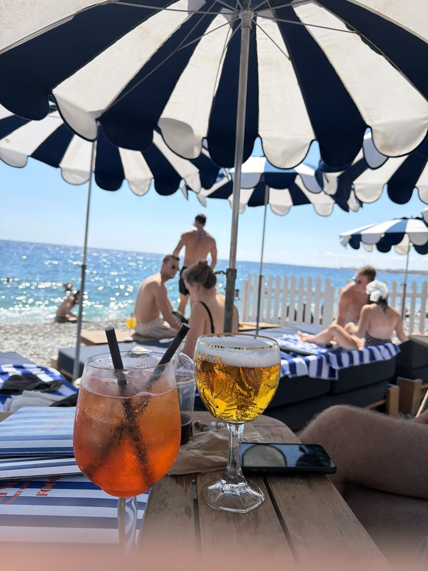 Spritz and beer on a table at a beach club with striped umbrellas and the sea behind