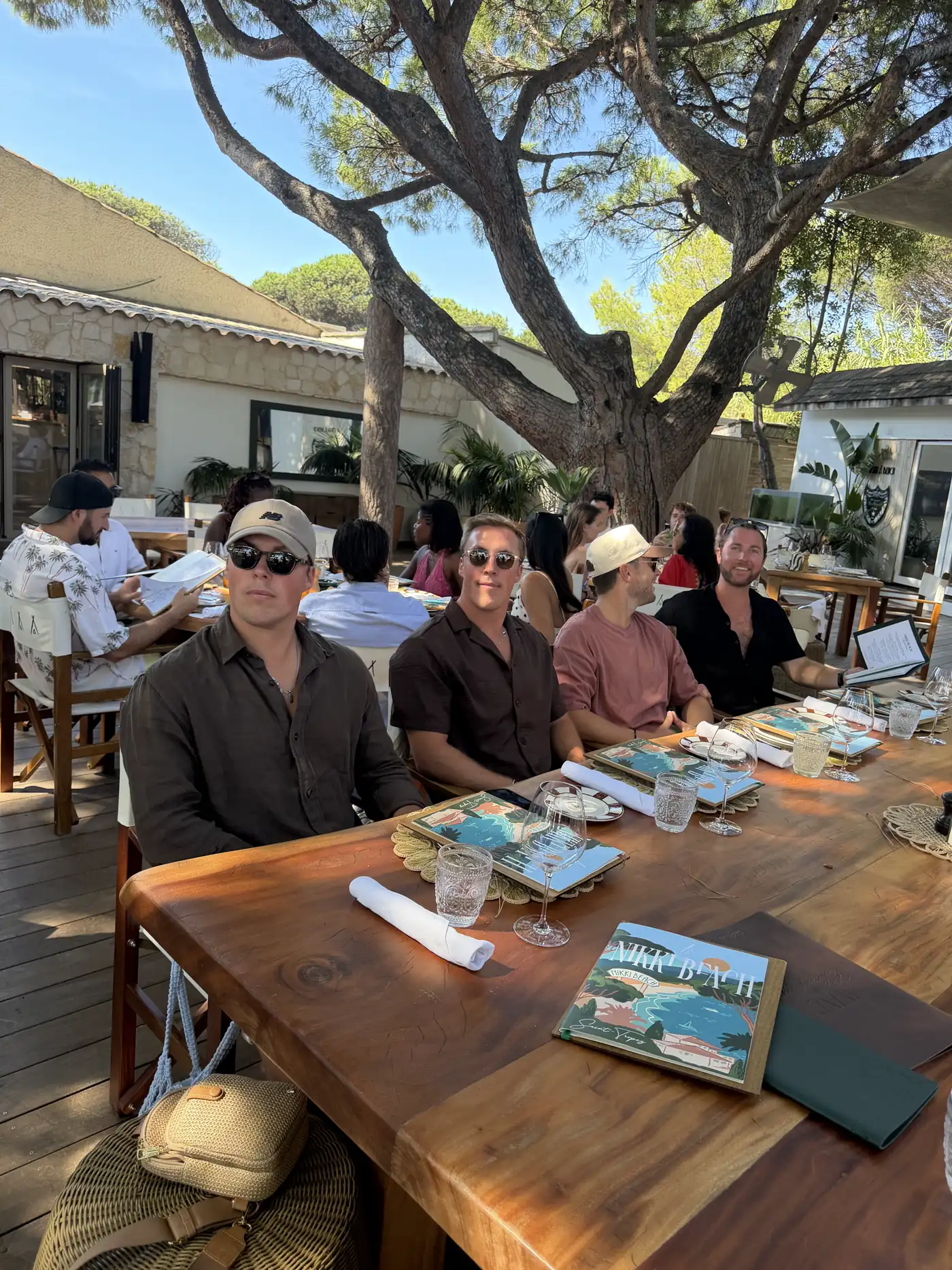 Friends at a long wooden lunch table under trees at a beach club