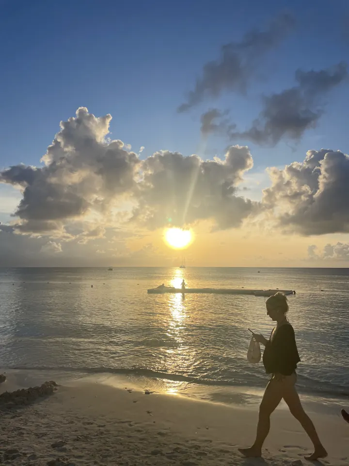 The sun sets over the sea behind clouds while a person walks along the beach