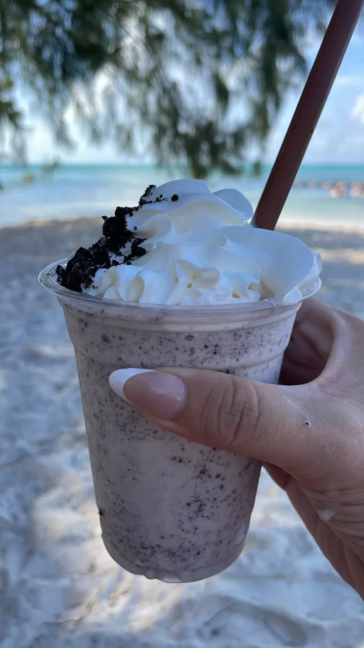 A hand holds a frozen cookies-and-cream drink with a straw on the beach