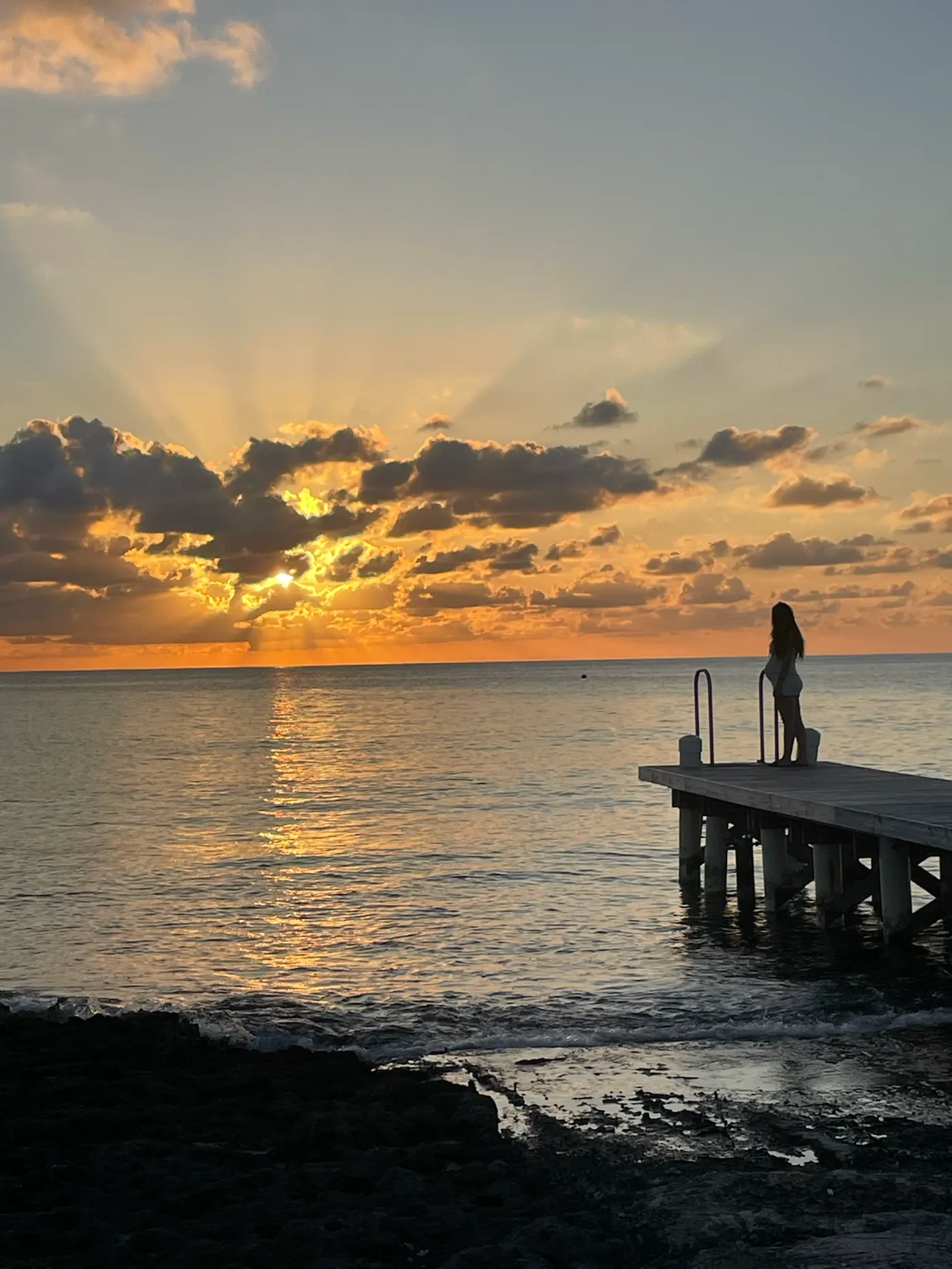 A person stands on a small pier at sunset over the sea