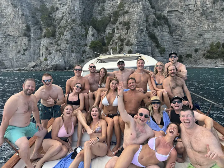 Big group of friends in swimwear on a boat below cliffs