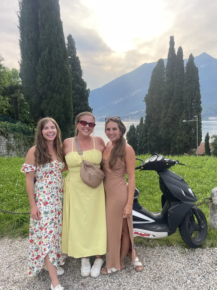 Three women in sundresses beside a scooter with cypress trees and lake