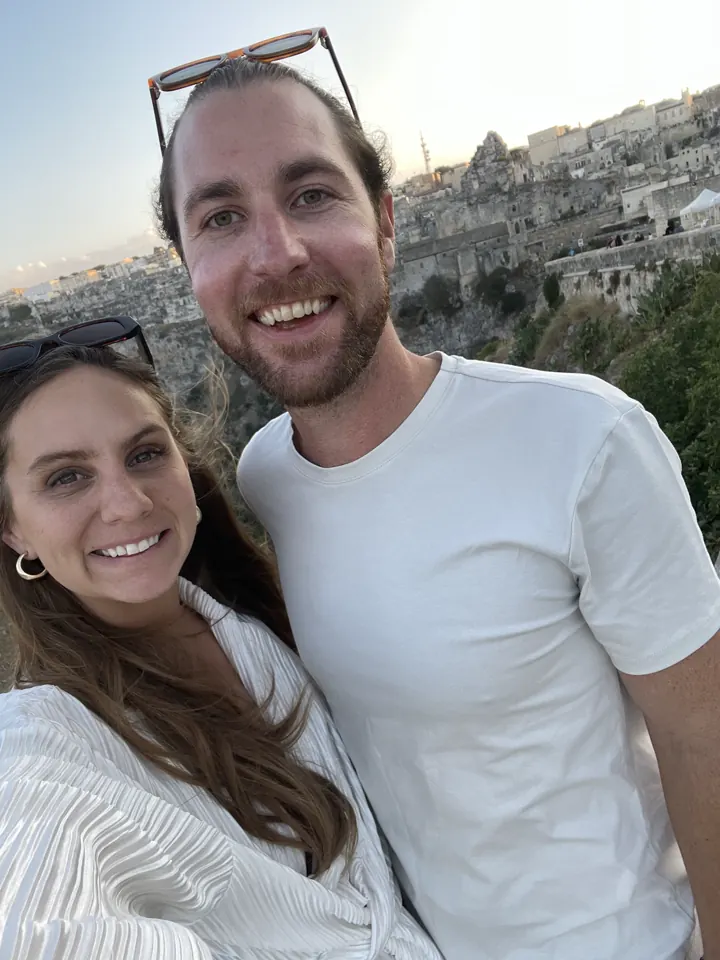 Colin and Angela smiling in a selfie with Matera's stone houses behind