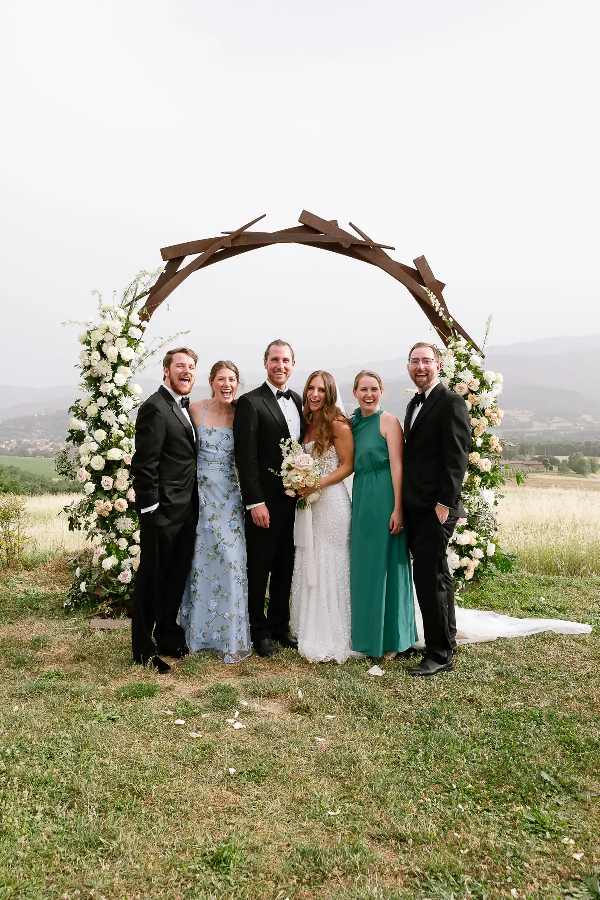 Wedding party in formal wear under a white floral arch on a lawn