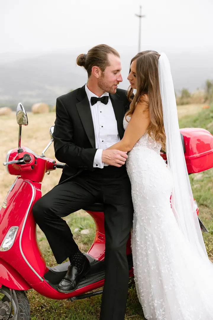 Colin in a tuxedo and Angela in her wedding gown posing with a red Vespa