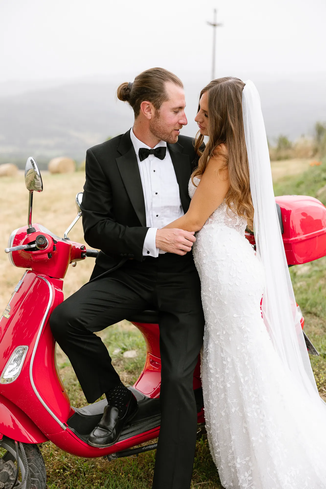 Colin in a tuxedo and Angela in her wedding gown posing with a red Vespa