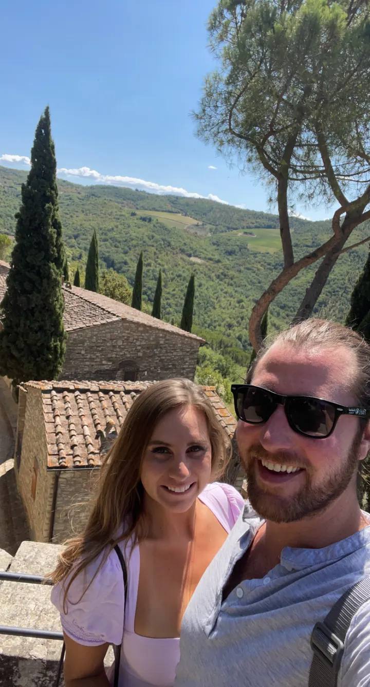A couple taking a selfie with cypress trees and green hills behind them