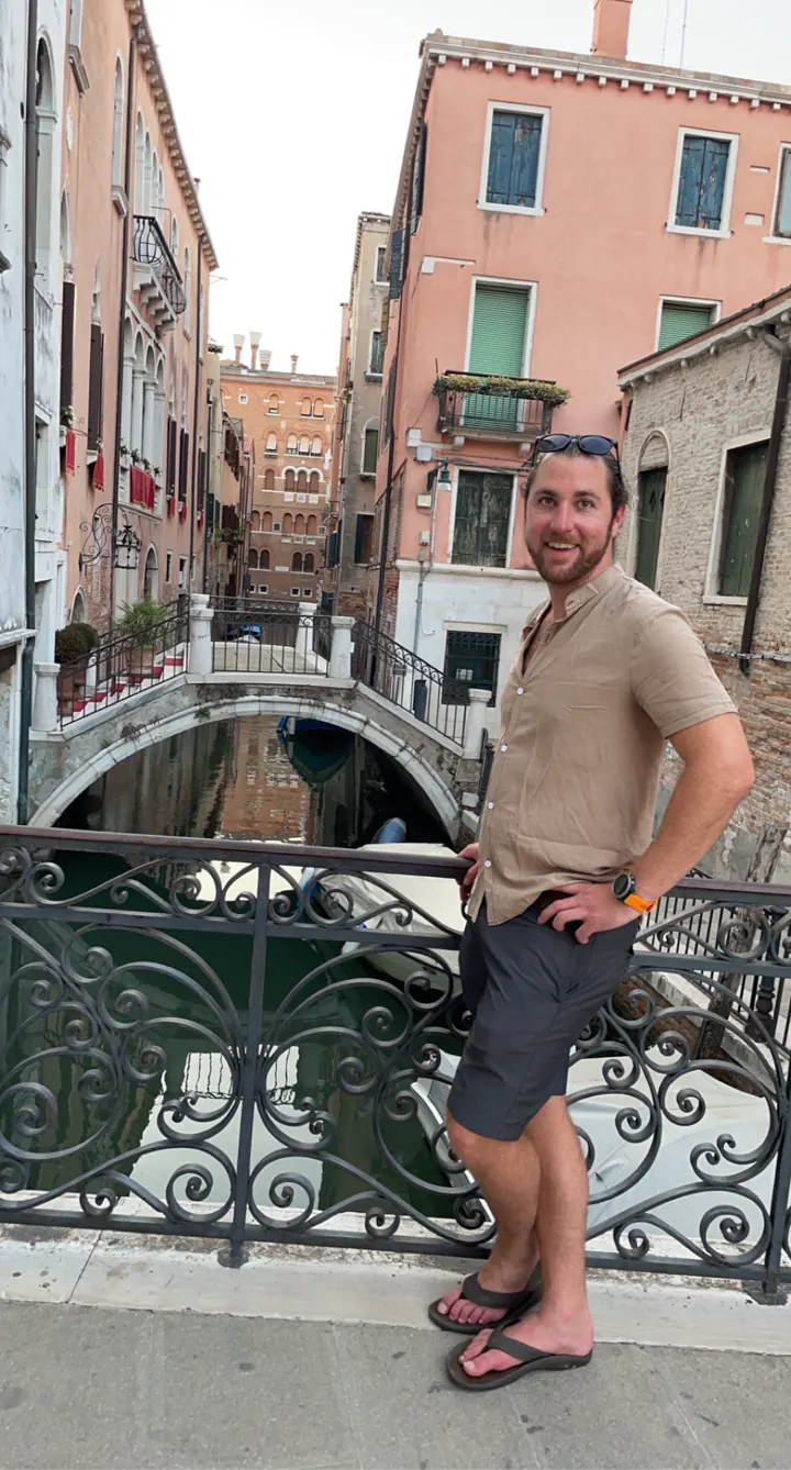 A man standing on a small canal bridge with an iron railing and pink buildings