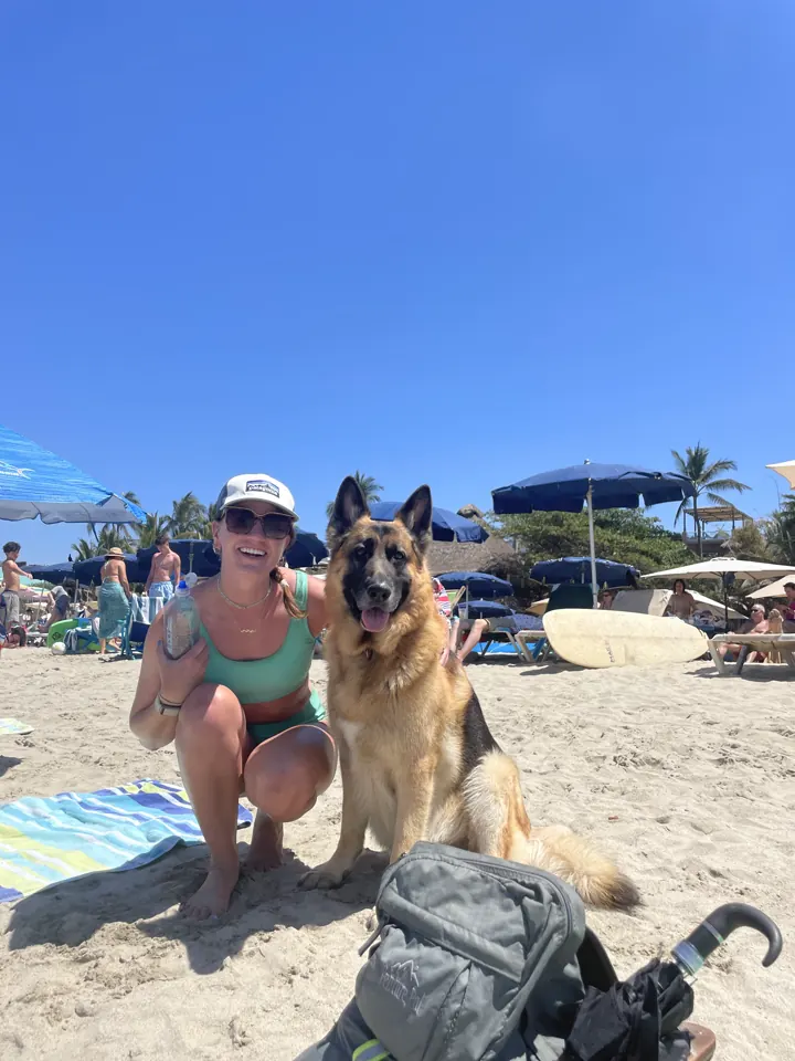 Angela kneeling on the beach with Jack, our German Shepherd, under colorful umbrellas
