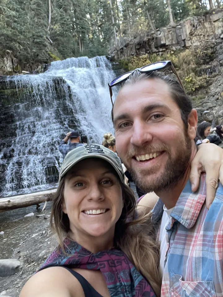 Angela and Colin smiling in a selfie in front of a cascading waterfall