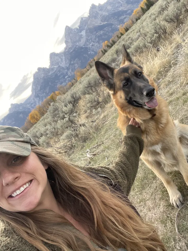 Angela taking a selfie with Jack, our German Shepherd, on a hillside with mountains behind