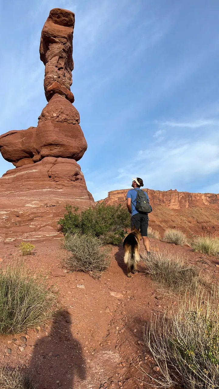 A tall balanced-rock hoodoo with a hiker standing nearby under a blue sky