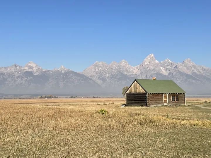 A small cabin with a green roof on a golden prairie beneath hazy mountains