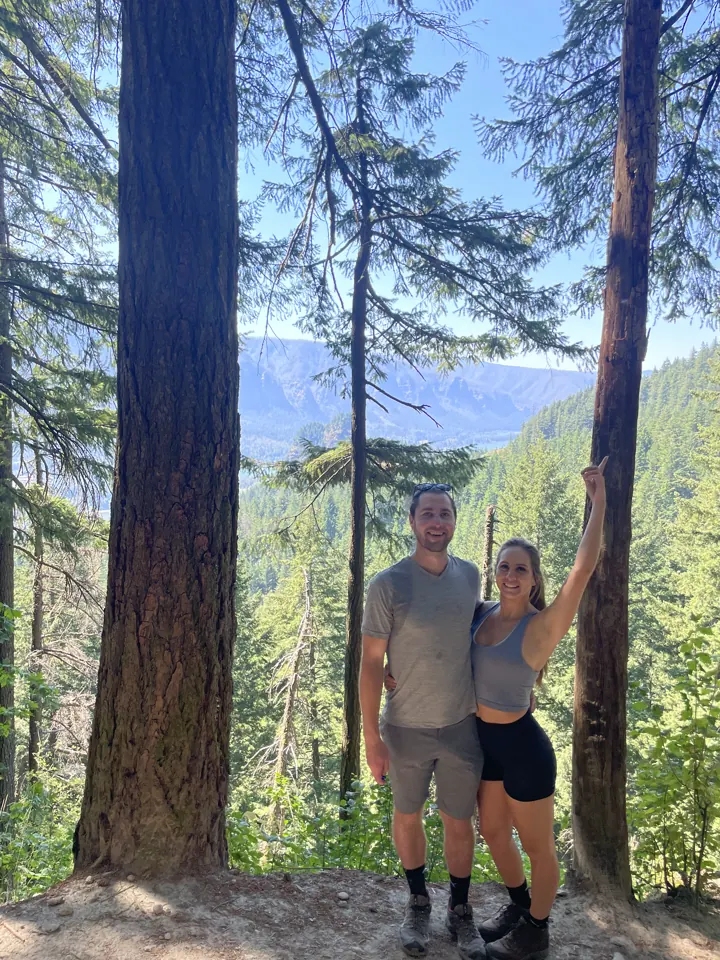 A couple standing among tall evergreen trees on a hike