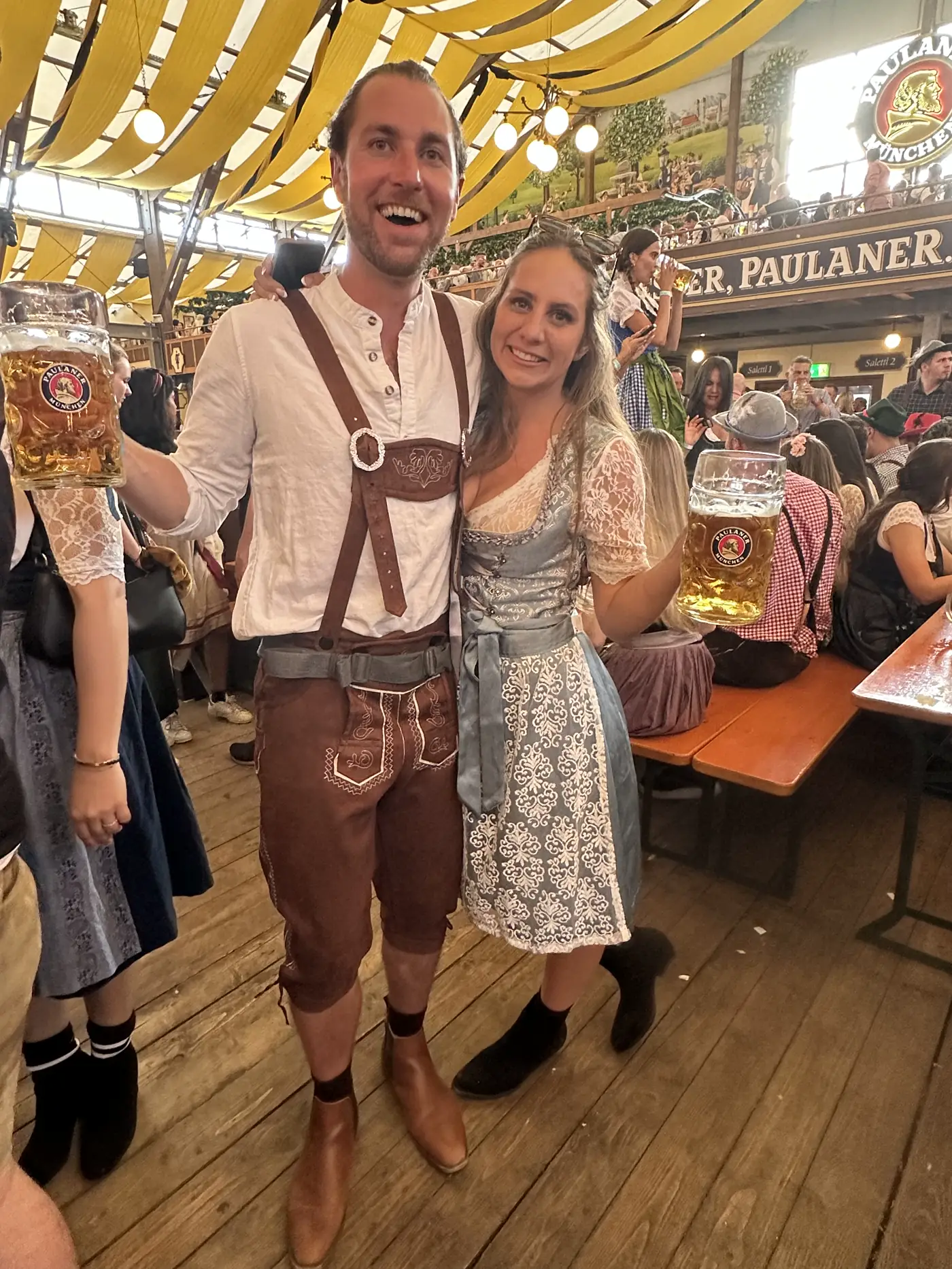 Colin in lederhosen and Angela in a dirndl holding beers in a beer tent