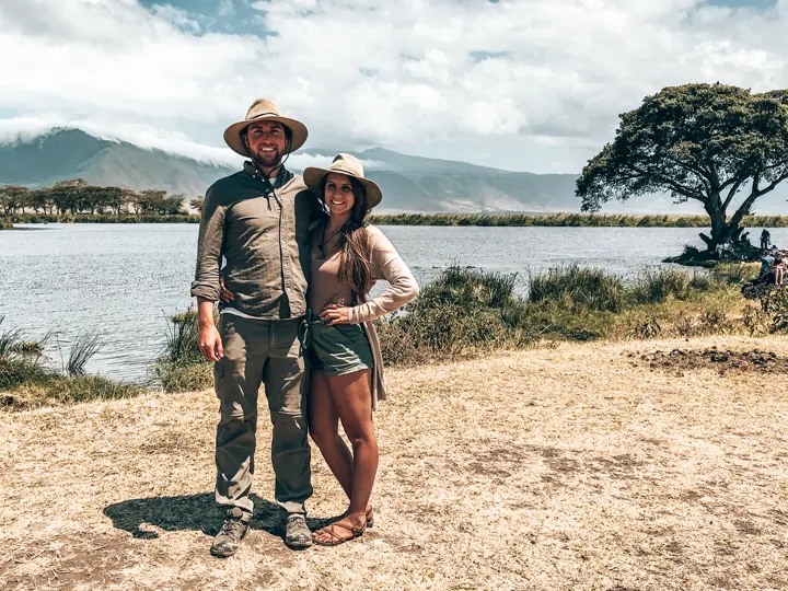 A couple in safari hats standing by a lake with an acacia tree behind