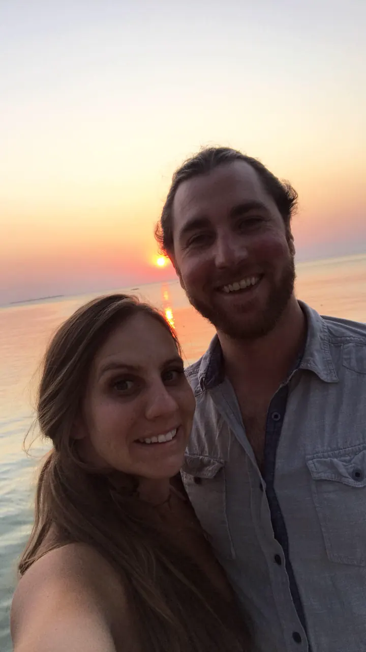 A couple taking a selfie with an orange sunset over the sea behind them
