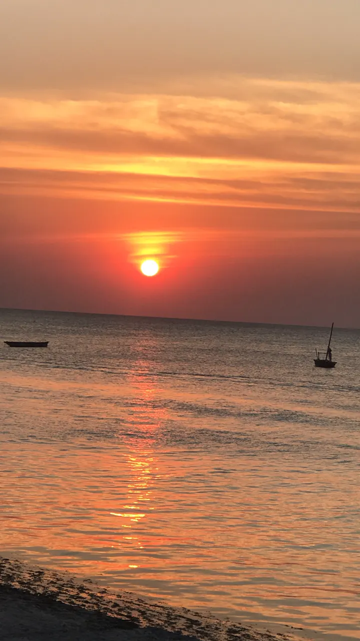 An orange sun setting over the ocean with a small dhow on the horizon
